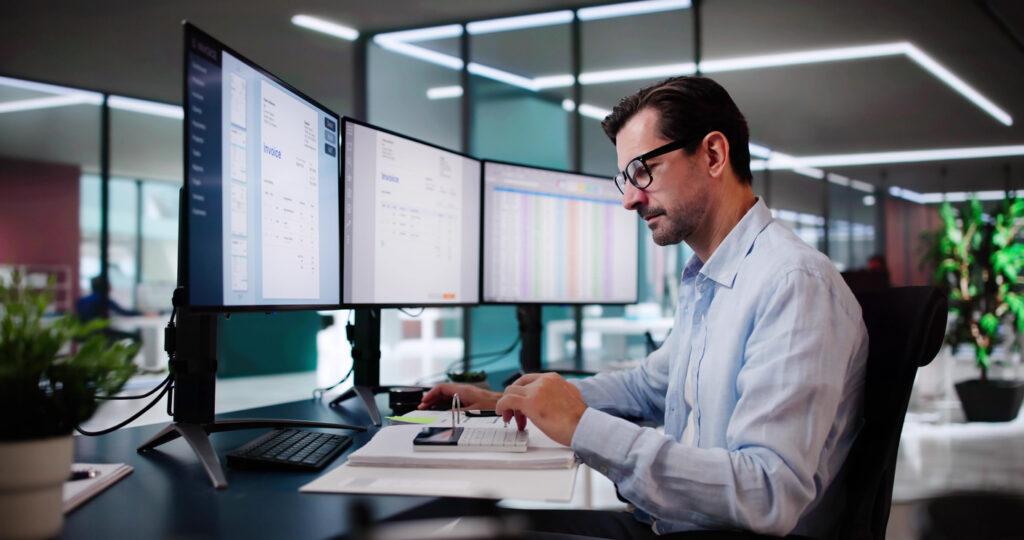 Profissional em escritório moderno analisando dados em monitores duplos, representando processos financeiros automatizado em ambiente colaborativo.